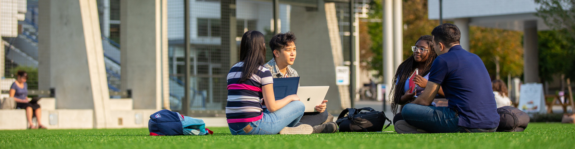 four students sitting outside on grass chatting holding laptops